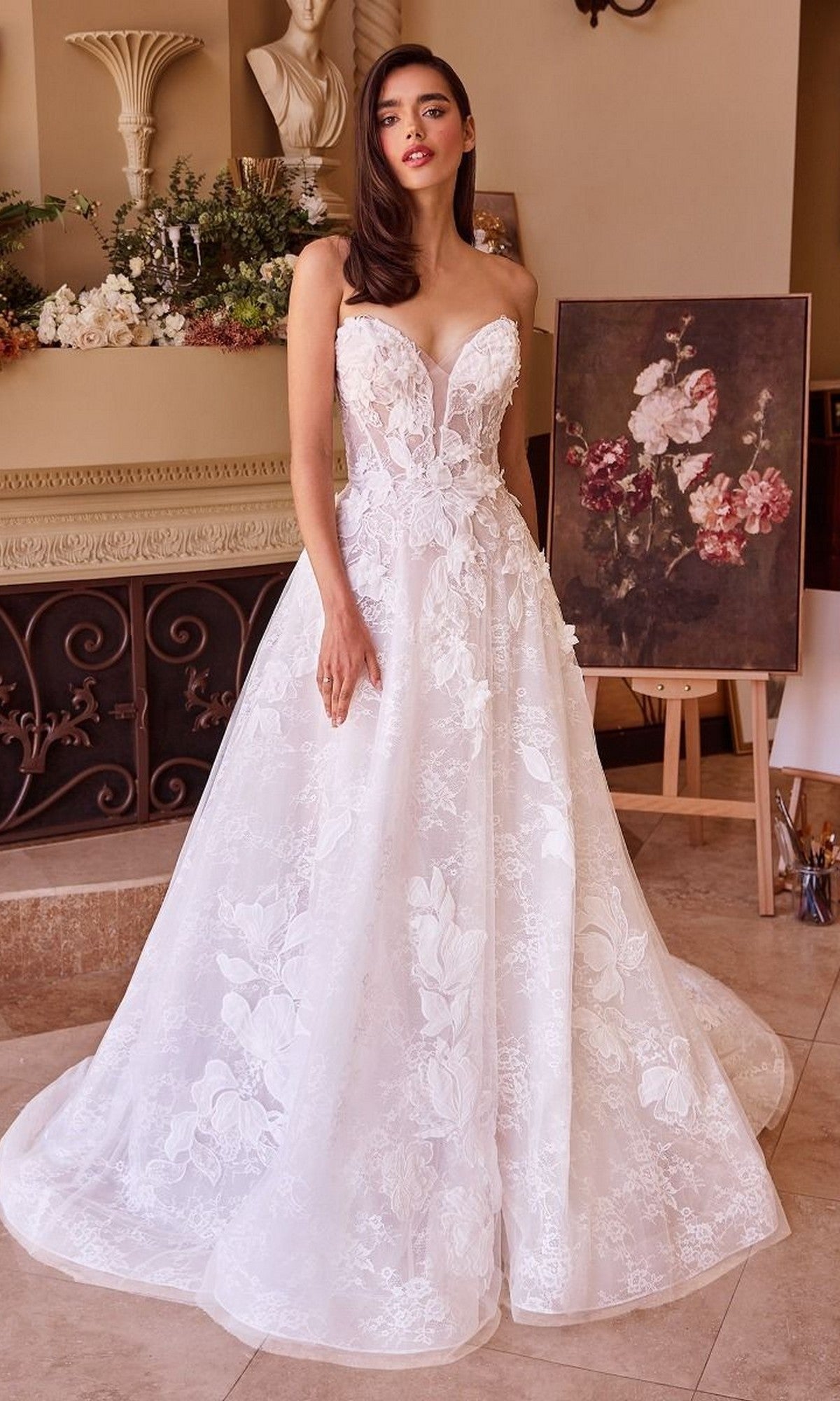 A woman stands indoors wearing the Andrea and Leo WL034 long wedding dress, a strapless gown adorned with floral lace appliques. Elegant floral arrangements, a classical bust, and a flower painting on an easel decorate the background.